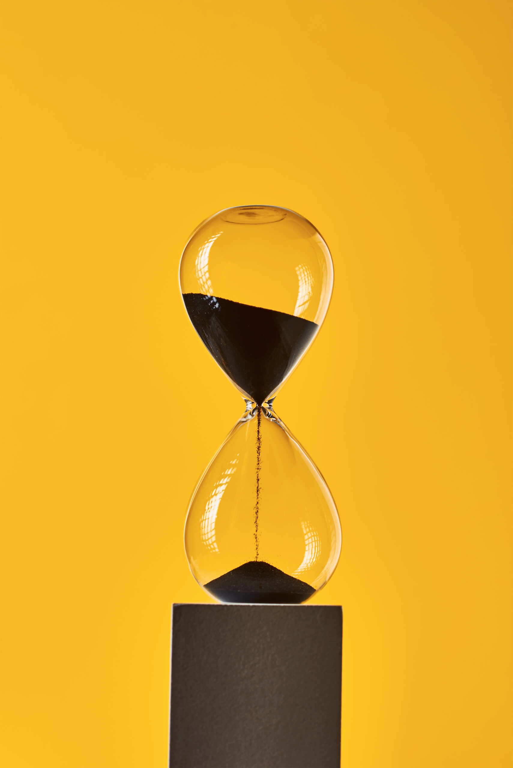 A clear glass hourglass with black sand flowing from the top bulb to the bottom, placed on a small black pedestal against a bright yellow background.