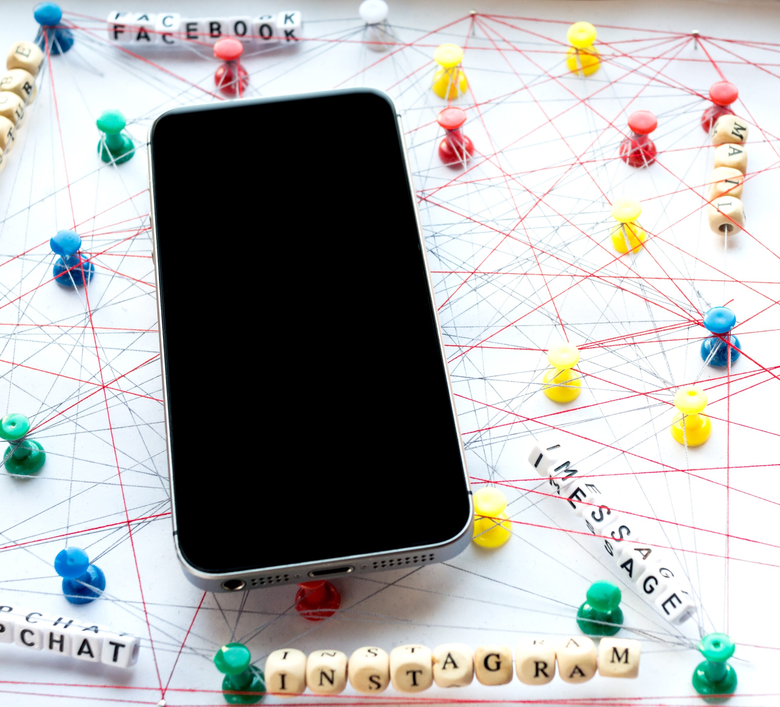 A smartphone lying face-up on a surface with push pins connected by red and blue strings, representing a network of social media connections. Wooden letter tiles spell out the names of apps such as Facebook, Instagram, Snapchat, and iMessage.